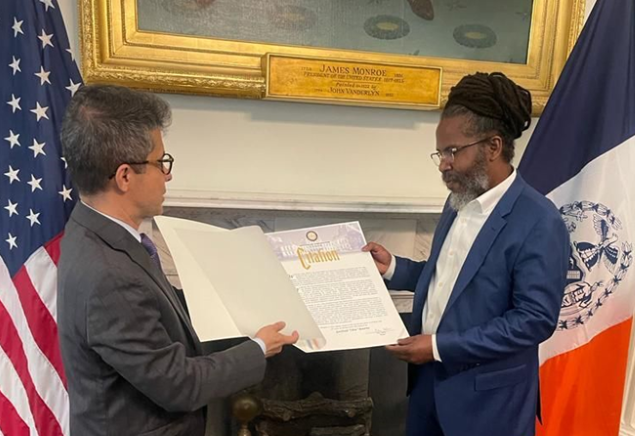 On behalf of New York City Mayor Eric Adams, Jose Bayona, executive director of the Office of Ethnic and Community Media presents Bourne with a citation.